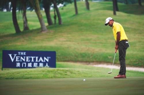 A man wearing a yellow t-shirt and olive green trousers and white hat plays golf in Macau.