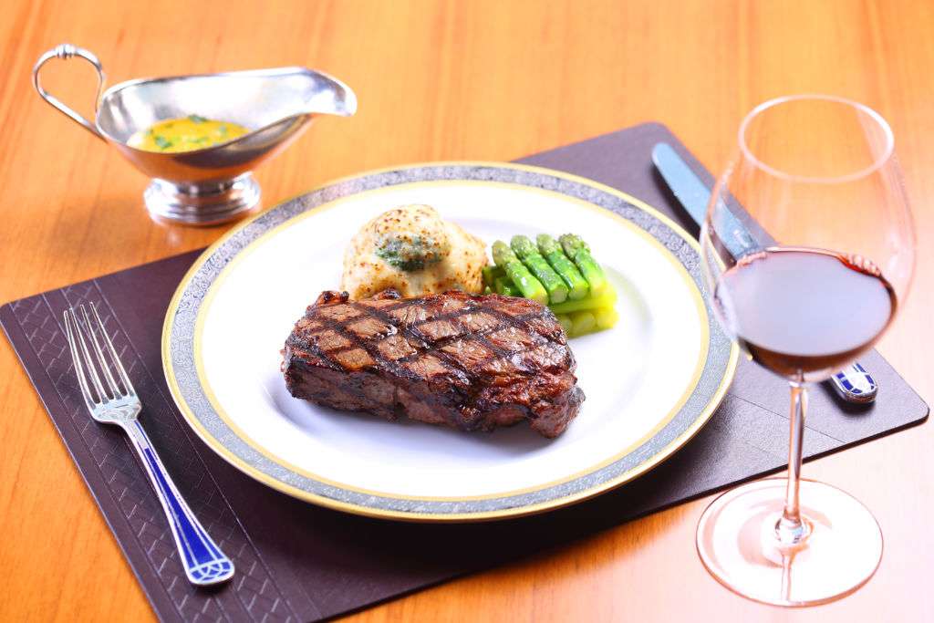 Steak with puree and asparagus on a plate mat, glass of red wine and gravy boat on a wooden table