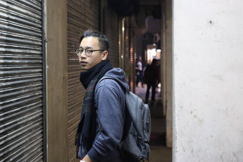 A young man wearing glasses, sweatshirt, scarf, and backpack poses in Macau alleyway at night. 