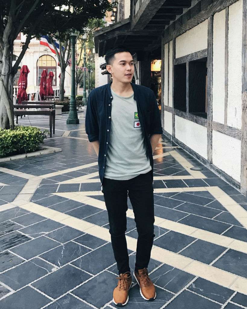 A young man poses outside on the street in Macau during the daylight, wearing a grey t-shirt, dark overshirt, dark trousers, and light brown shoes.
