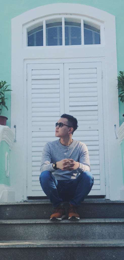 A young man poses outside in Macau during the daylight, wearing sunglasses, a light grey top, jeans, and light brown shoes.