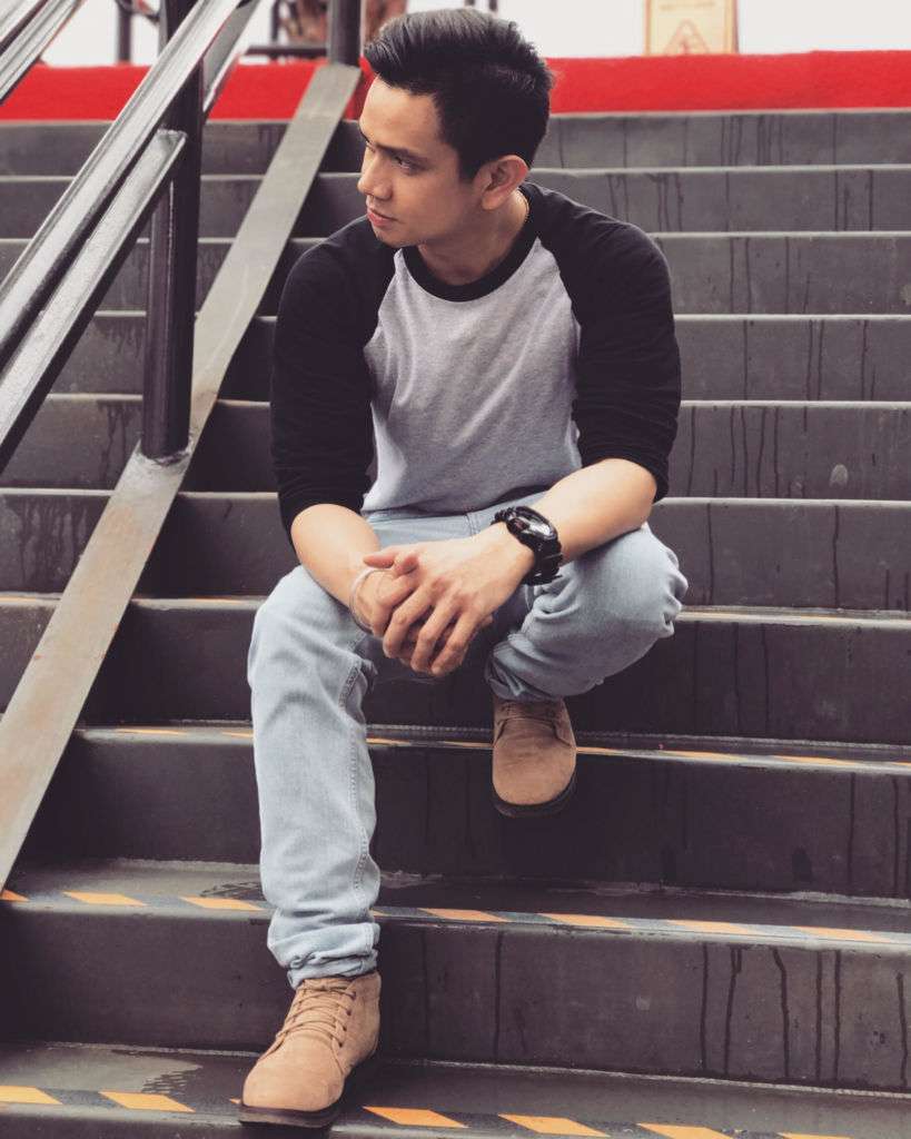 Louis Quizon sits outside on steps in Macau, wearing a shirt, light blue jeans, and boots.