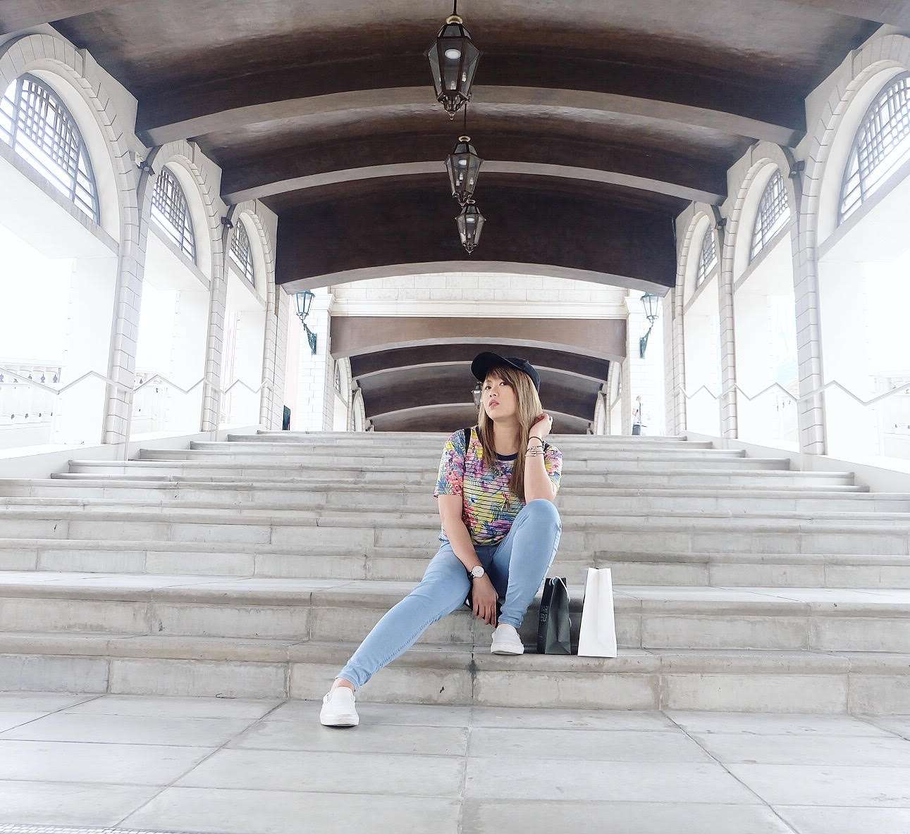 A girl poses outside in Macau on some steps, wearing a dark hat, multicolored top, jeans, and white shoes.