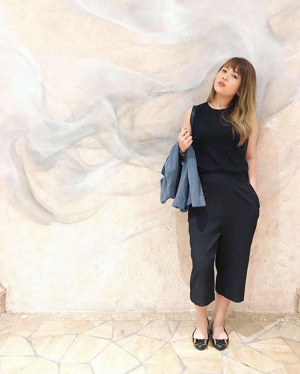 A girl in Macau poses against a wall wearing a black top and slacks. 