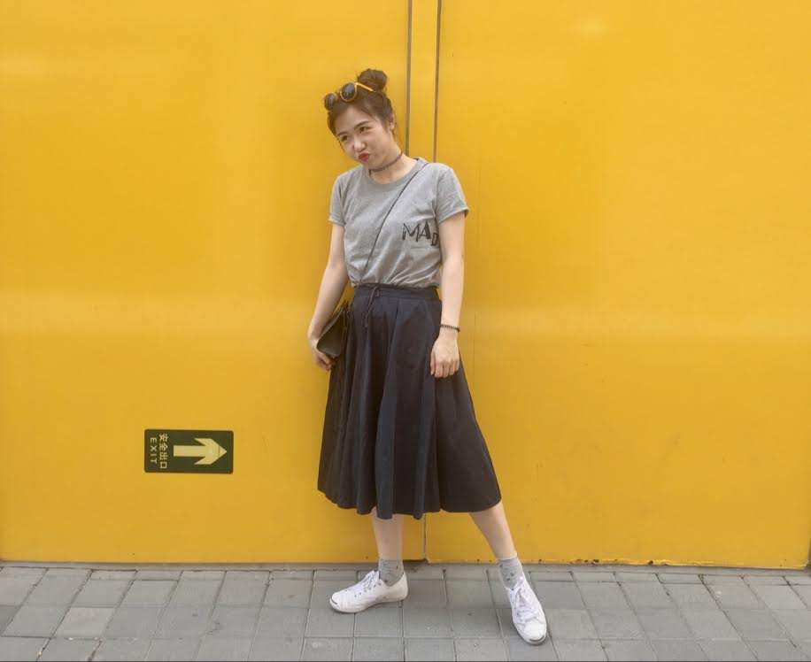 Sueie Che, a girl in Macau, poses in front of yellow backdrop outside on the street, wearing a long dark colored skirt and a grey t-shirt. 