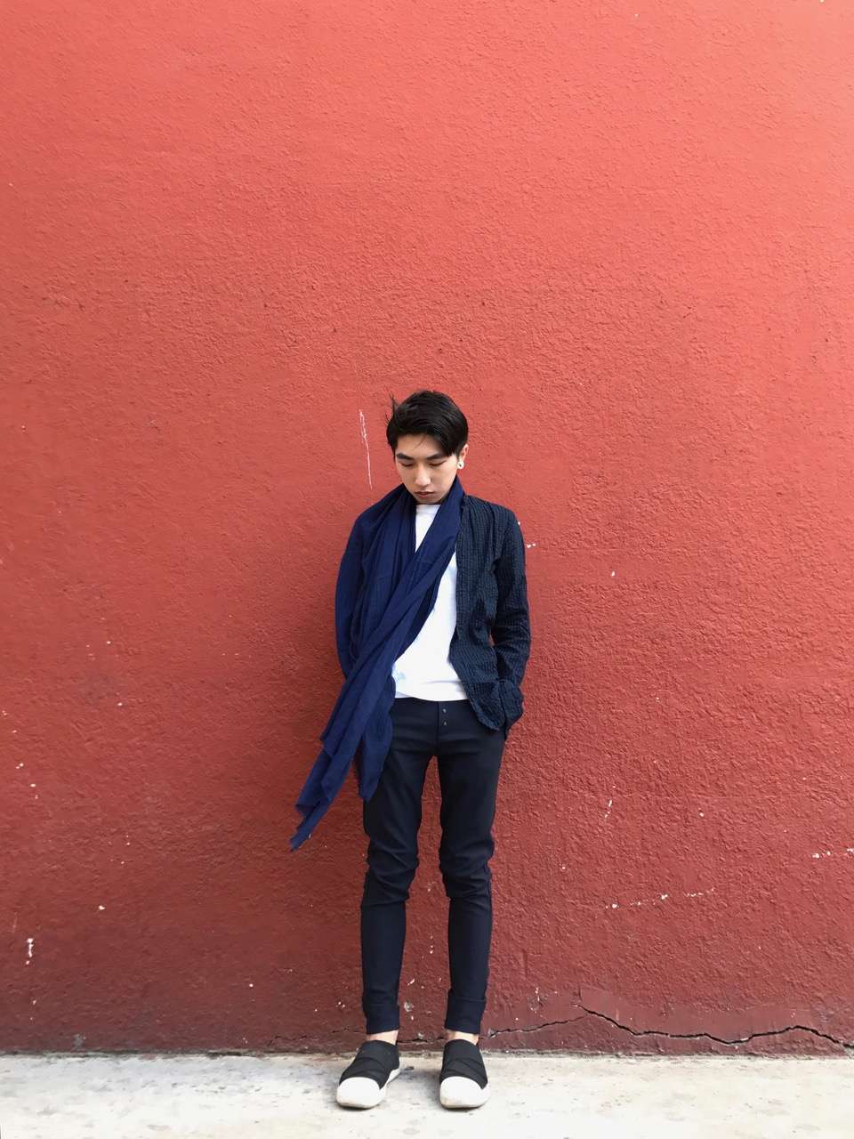 A young man poses outside in Macau against a red wall, wearing a dark top and white shirt with long blue scarf. 