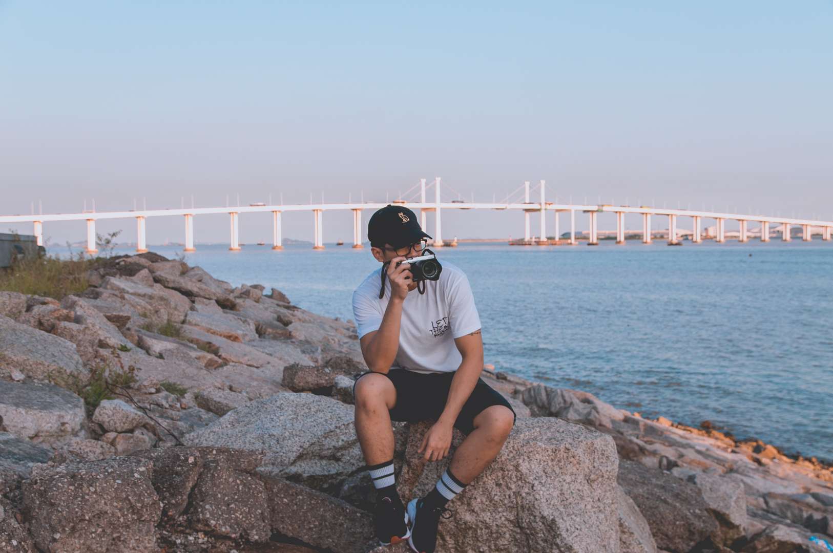 Lance Relucio sits on rocks near the water with Taipa bridge in the background. 