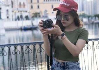 Girl outside in Macau wearing red hat, green top, denim shorts, and holding a camera.