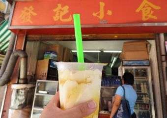 A hand holding up a drink in front of the sign for Fat Kei Siu Sik shop in Macau