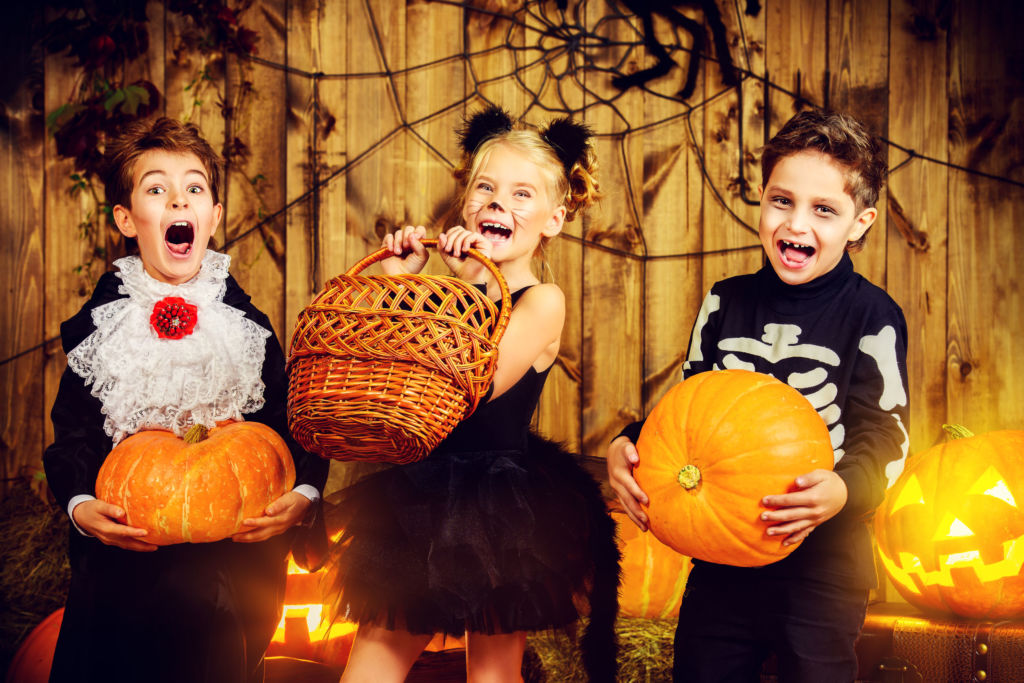 Two boys and a girl in Halloween costumes while holding pumpkins and a basket