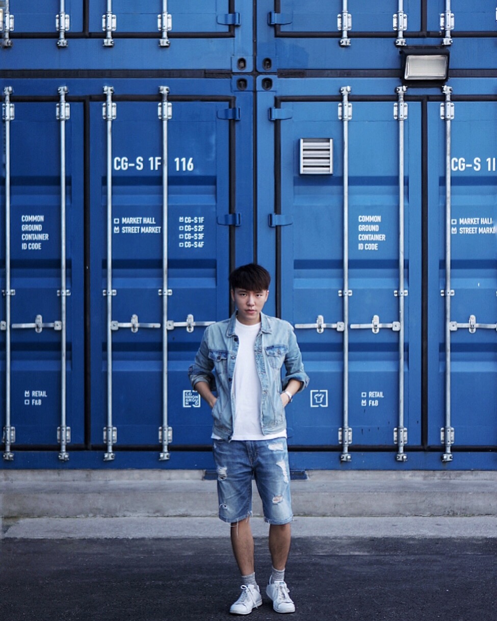 A young man wearing a denim jacket, white t-shirt, and denim shorts poses in front of a blue background in Macau.