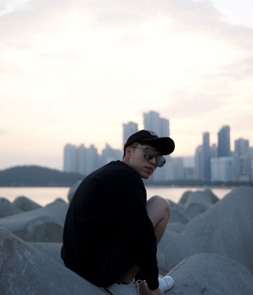 A young man wearing a dark hat and sunglasses sits on a rock with the Macau cityscape in the background.