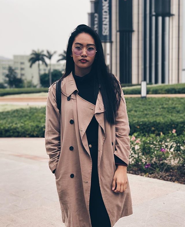 A young woman poses standing outside in Macau, wearing large wire-rimmed glasses and a long light brown overcoat.