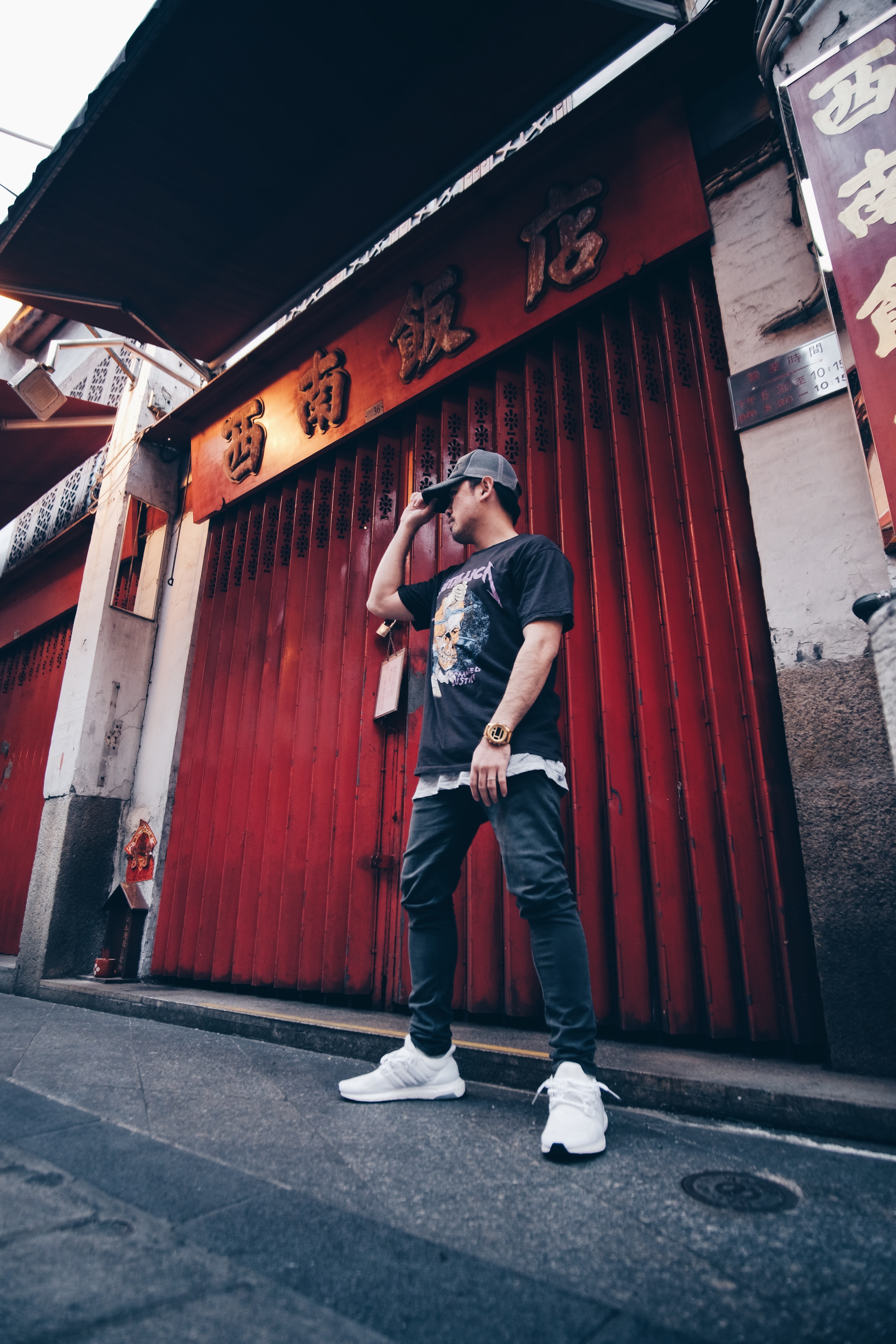 A young man wearing a hat, dark shirt and pants, and white shoes stands on the street in Macau.