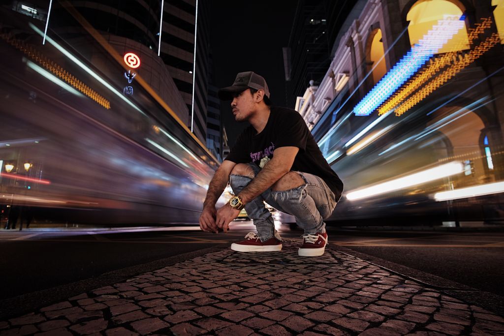 A young man wearing a hat and jeans kneels on the street in Macau