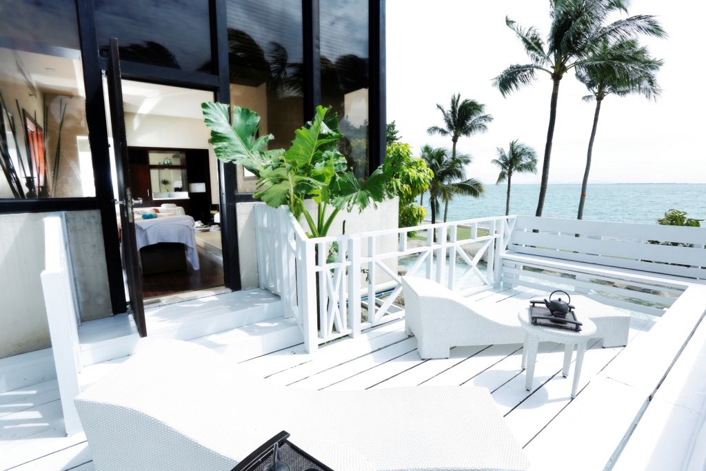 A beachside veranda painted white with matching white deck furniture. Palm trees in the background and distance. 