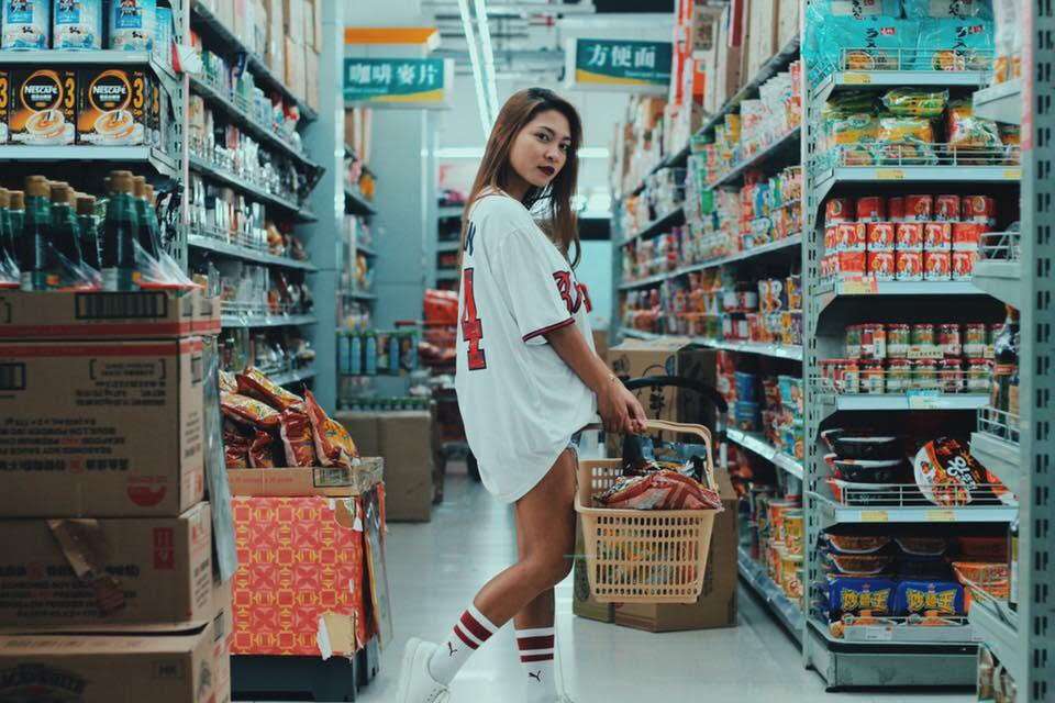 A young woman poses in a supermarket, holding a shopping basket.