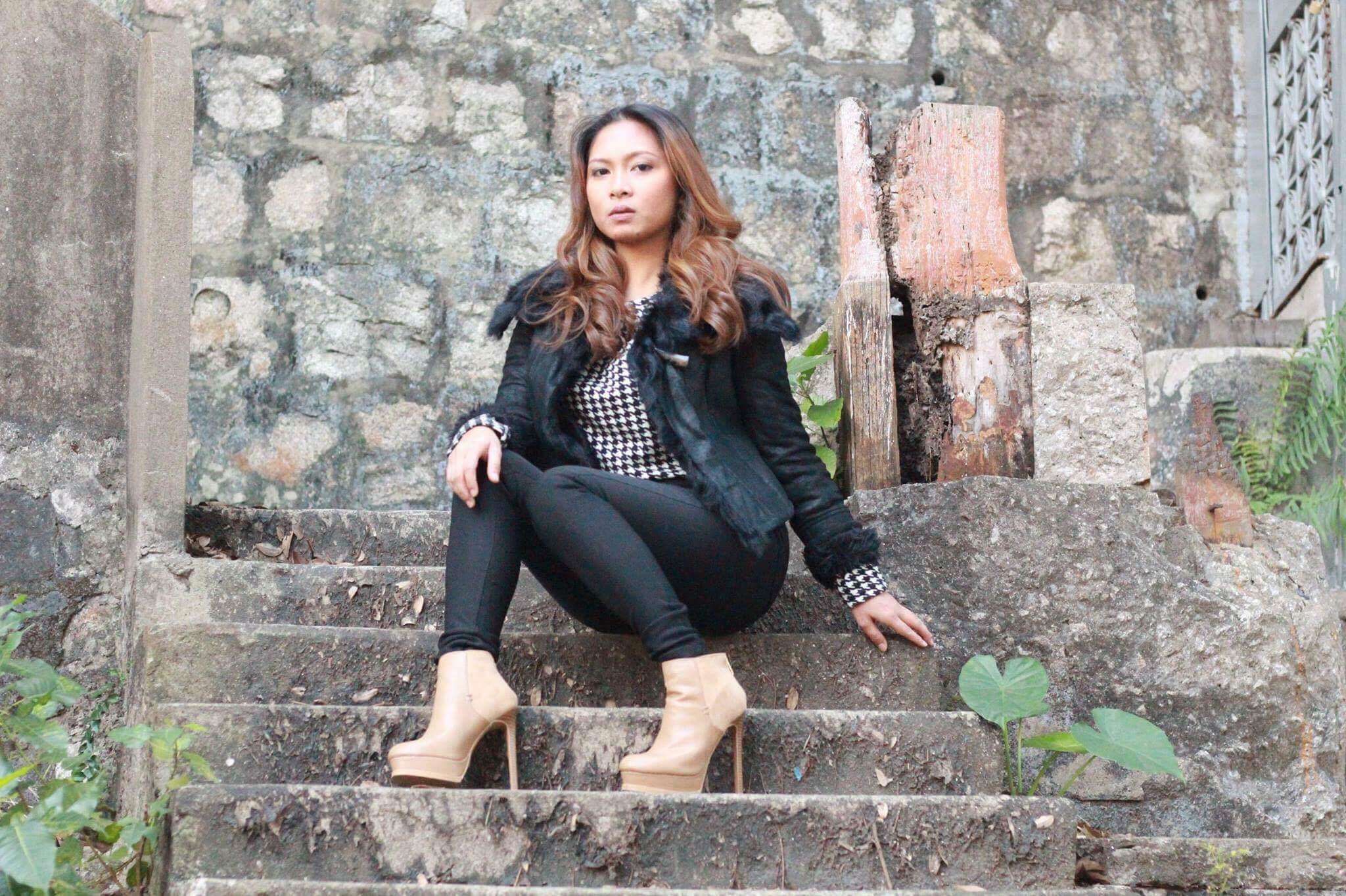 A young woman wearing a black jacket and black pants poses on some step outside in the daylight. 