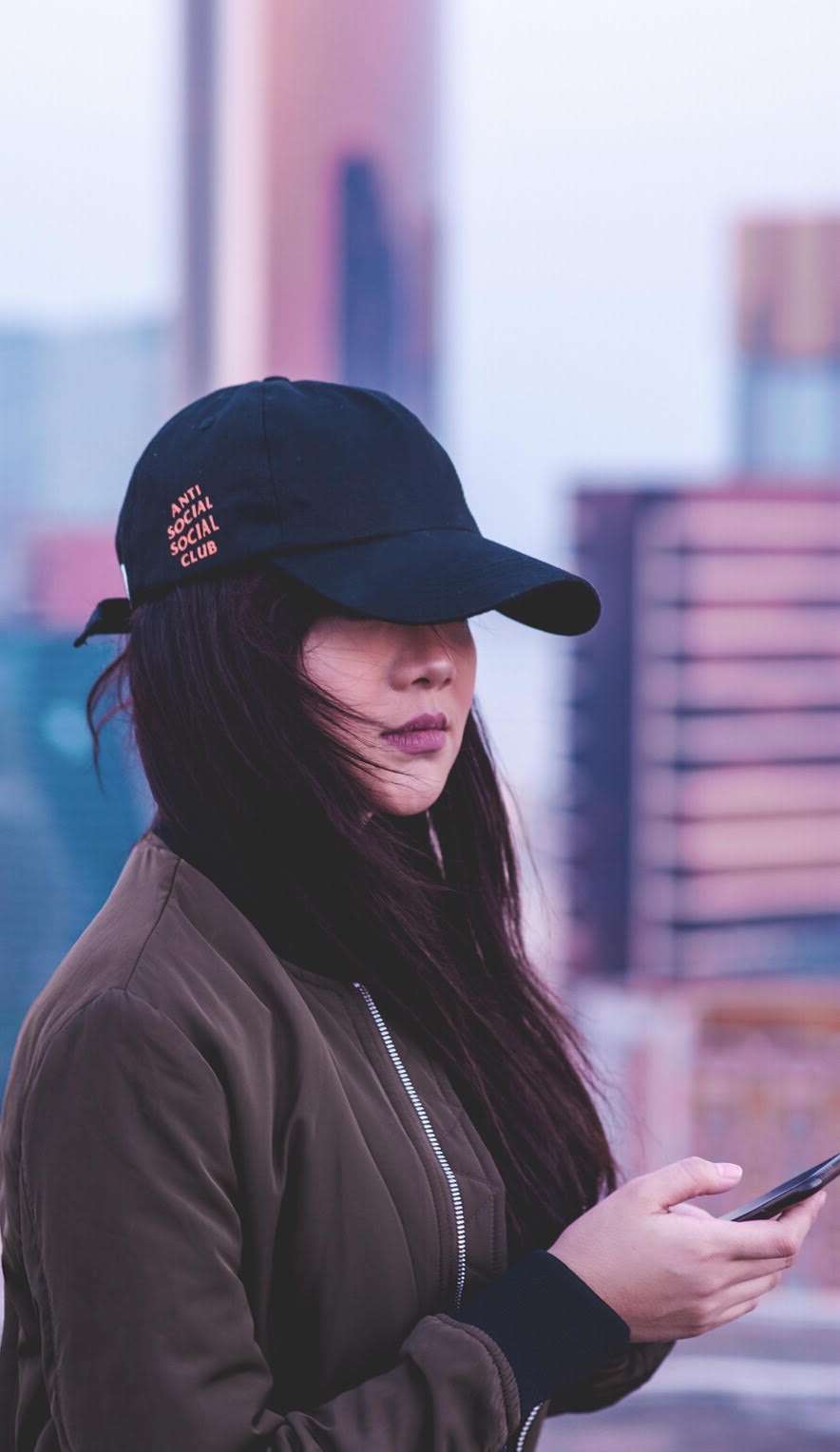 A young woman outside in Macau wearing a dark baseball cap, brown jacket, and holding a mobile phone.