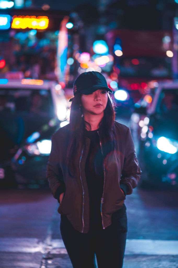 A young woman on the street at night wearing a baseball cap and dark outfit.